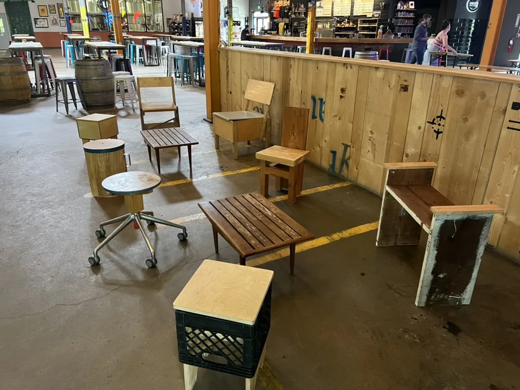Chairs and stools spread across the Union Craft Brewing taproom floor along a plank wall: a milk-crate stool with a plywood seat, a slatted bench, a stump, an office-chair base with a round wooden seat, and several plywood chairs