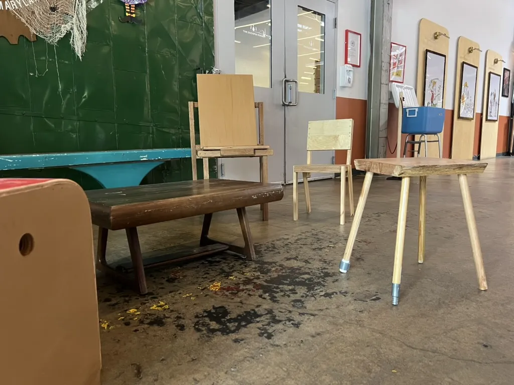 A low slatted table, a tall plywood chair, a slim pale chair and a three-legged stool on splayed dowel legs, in front of a green wall