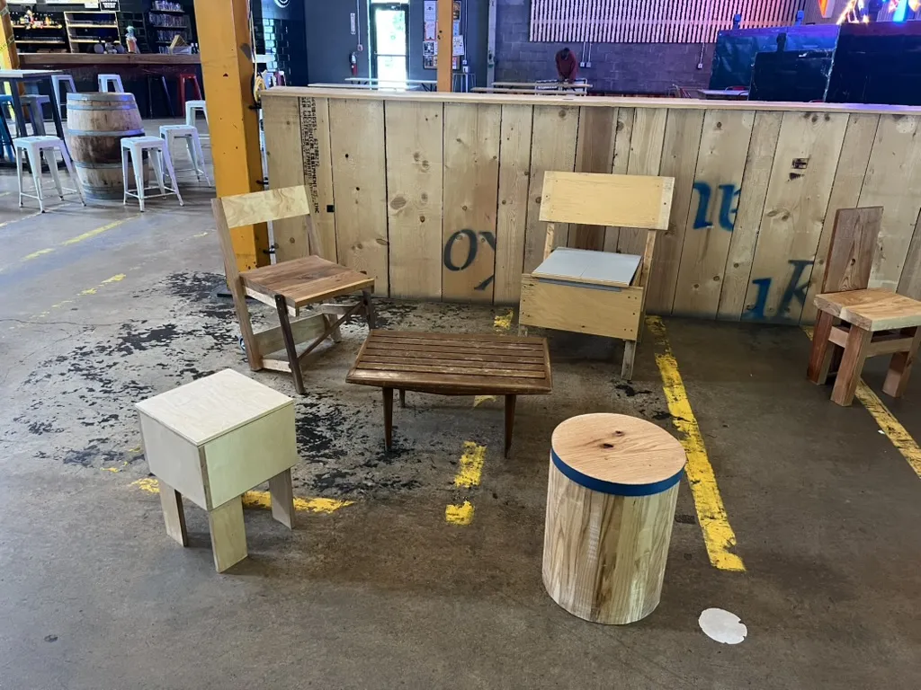 A white plywood box stool, a log stool banded in blue and two plywood chairs, one with a gray cushion, around a slatted table