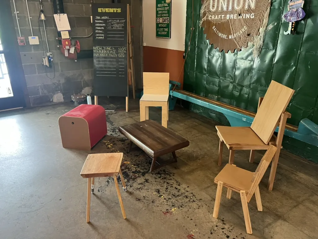 A red rounded stool, a low dark table, a tall plywood chair, and smaller pale wood chairs and stools beside the taproom’s events board