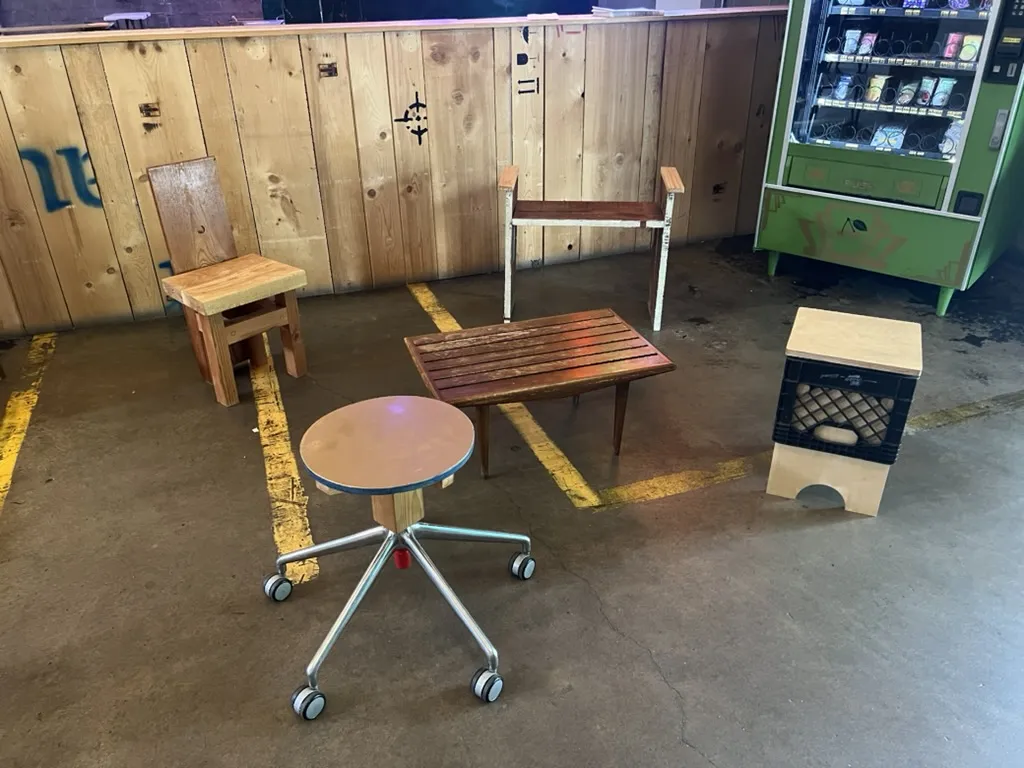 An office-chair base topped with a round wooden seat, a slatted table, a milk-crate stool and two chairs in front of the plank wall