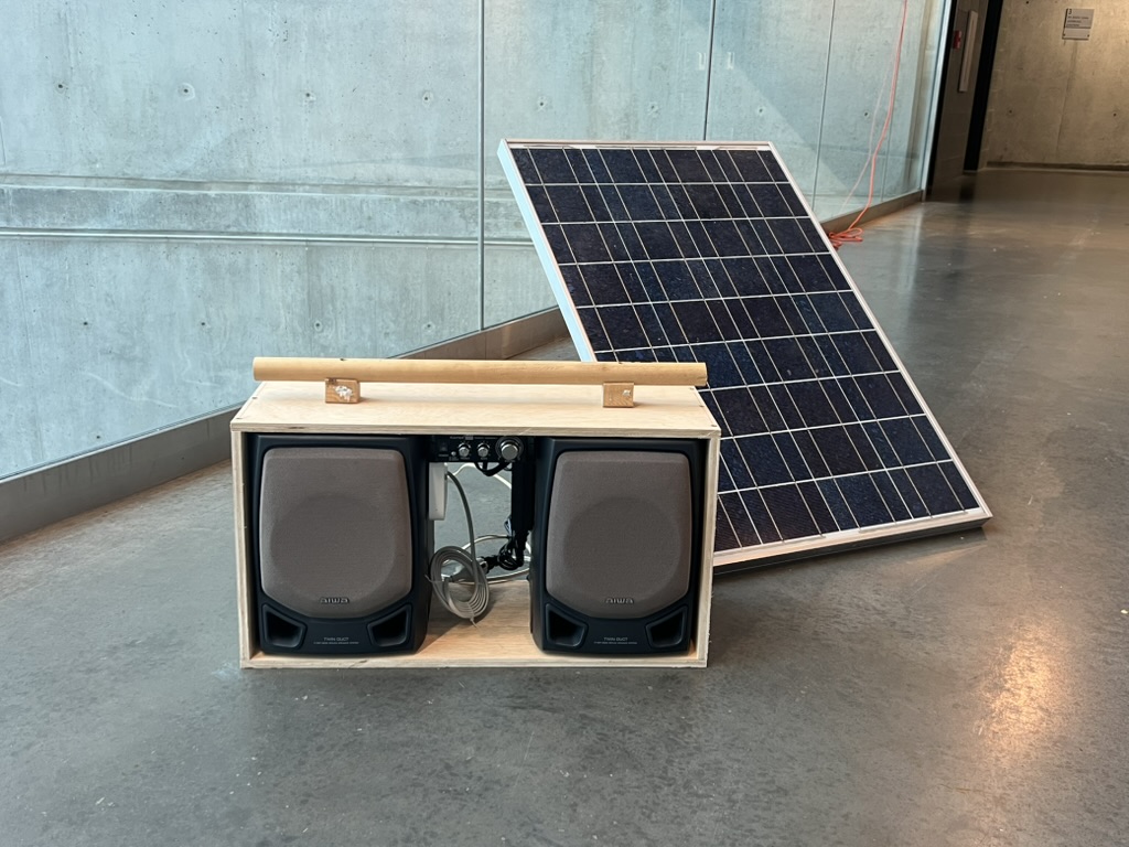 Finished Solarpunk Boombox next to the solar panel it charges from, photographed at an angle in a hallway
