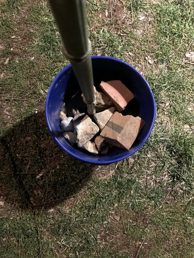 Looking down into a blue five-gallon bucket packed with broken brick and stone around the base of a bamboo pole, set on grass