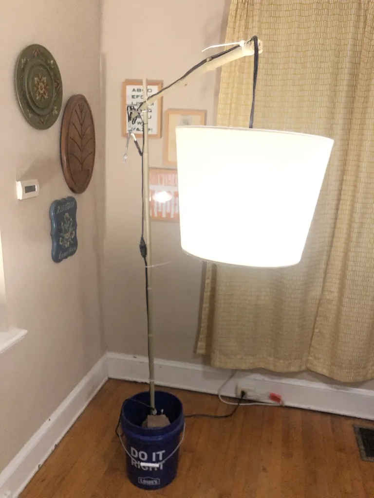 The first bucket lamp in a corner of a room: a bamboo pole in a blue bucket, its arm holding a glowing white drum shade